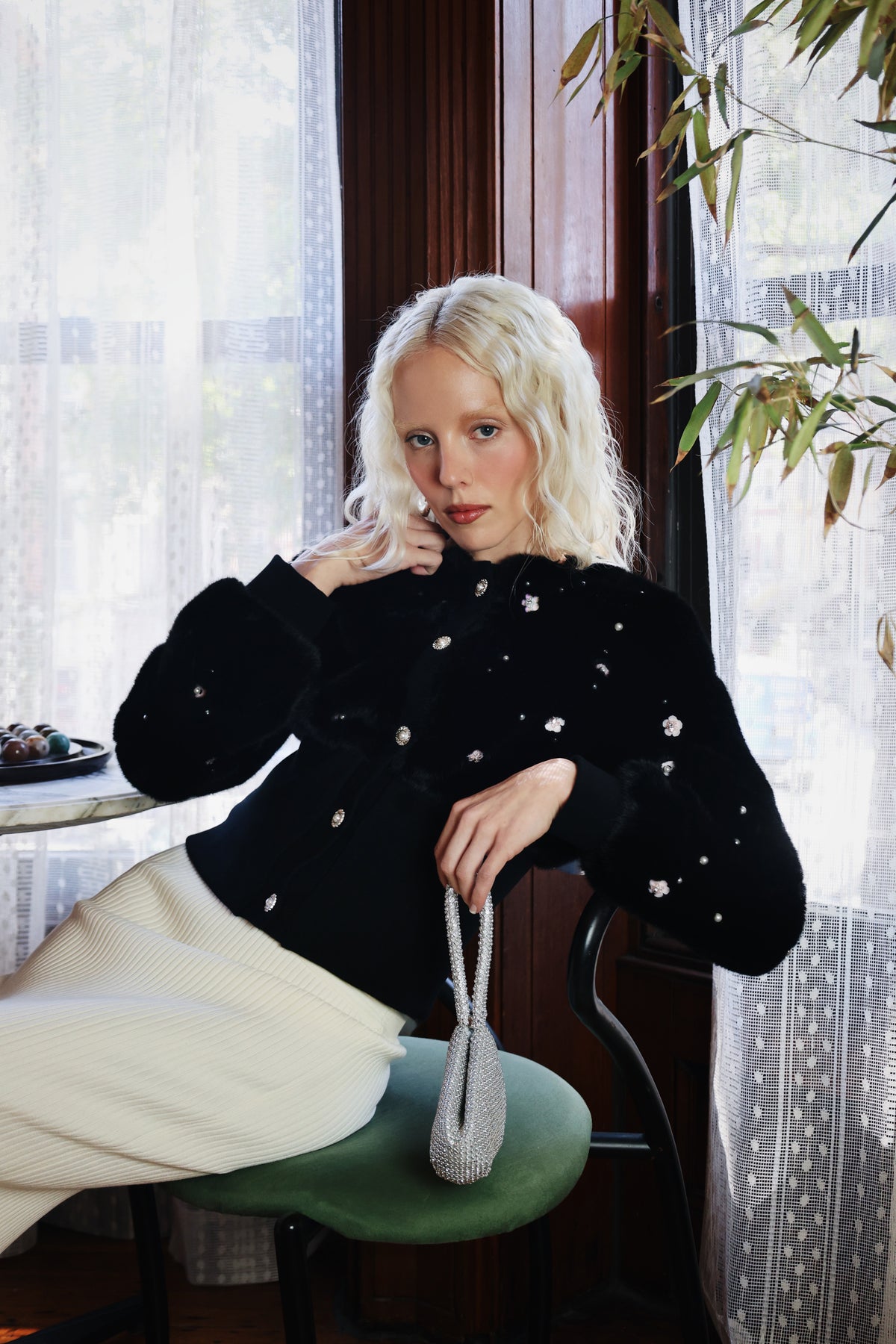 Model in the Pearle faux fur cardigan jacket and a cream skirt, seated by a lace-curtained window, Dollhouse AW'26 campaign