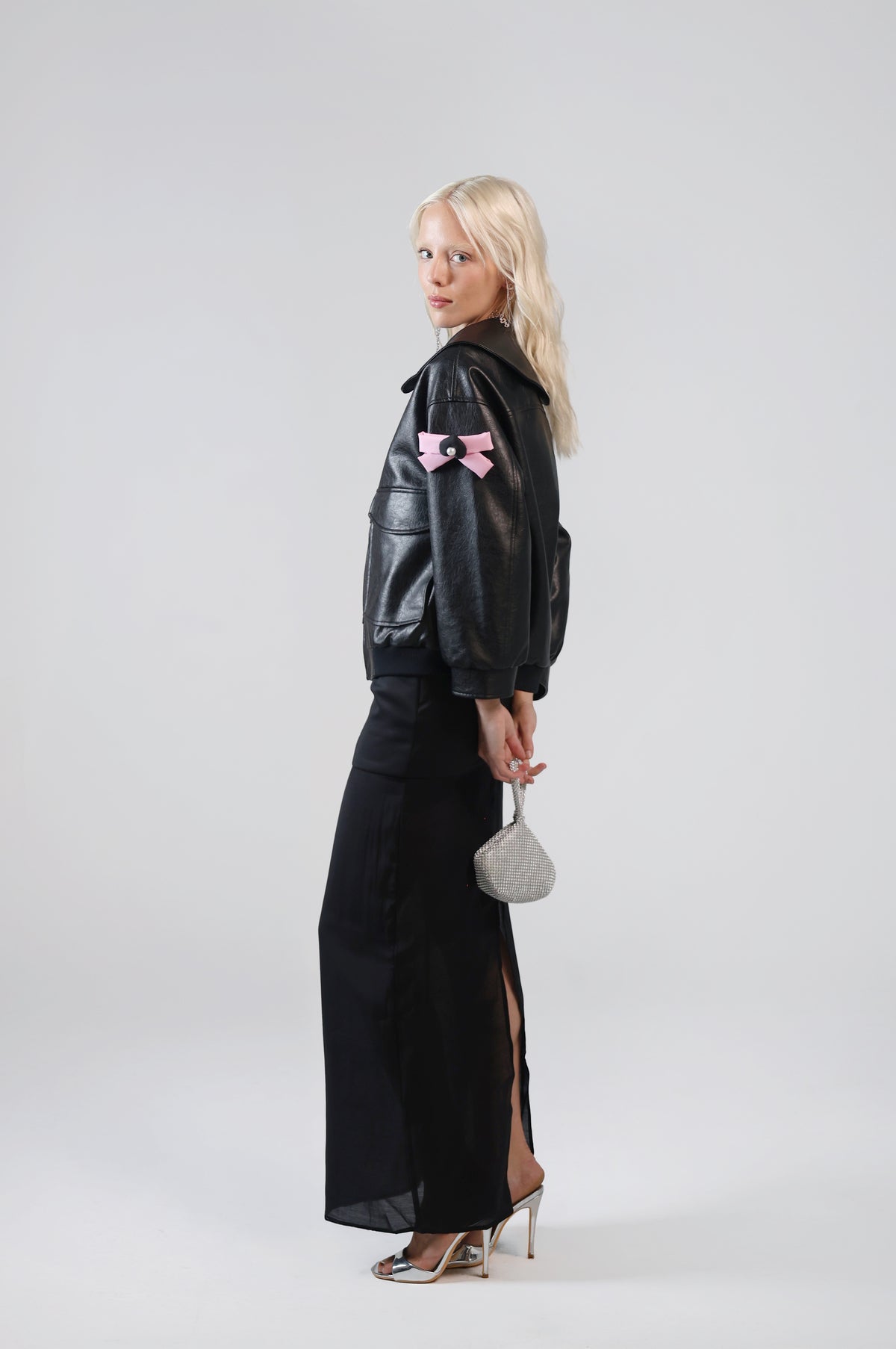 Jinnoir black faux leather jacket with pink bow charms on the sleeve, side view on model with a black maxi skirt