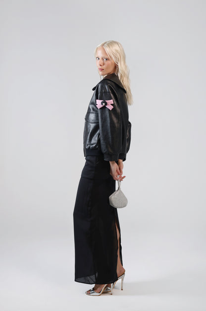 Jinnoir black faux leather jacket with pink bow charms on the sleeve, side view on model with a black maxi skirt